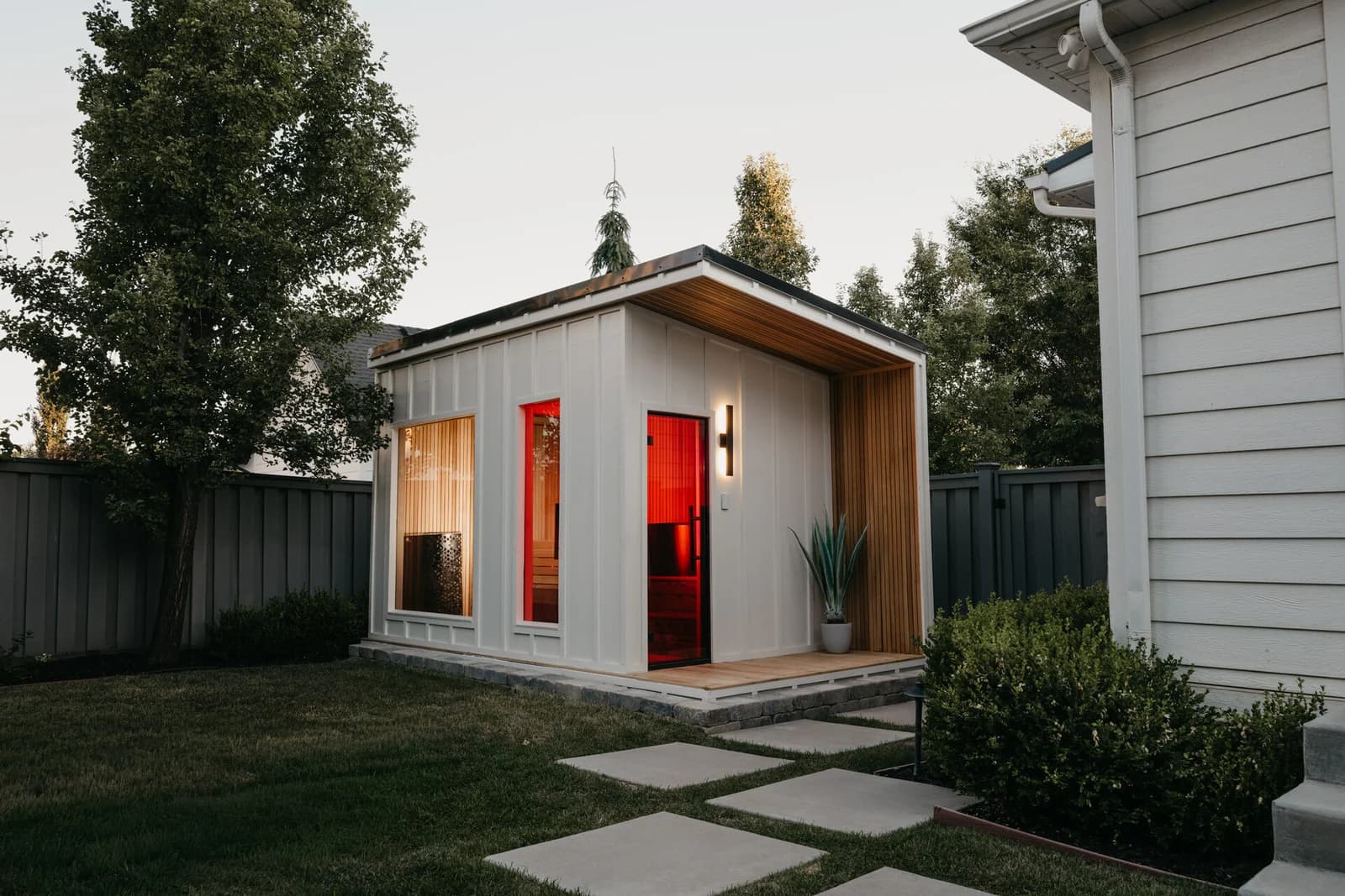 A timber backyard sauna illuminated by warm red interior light.